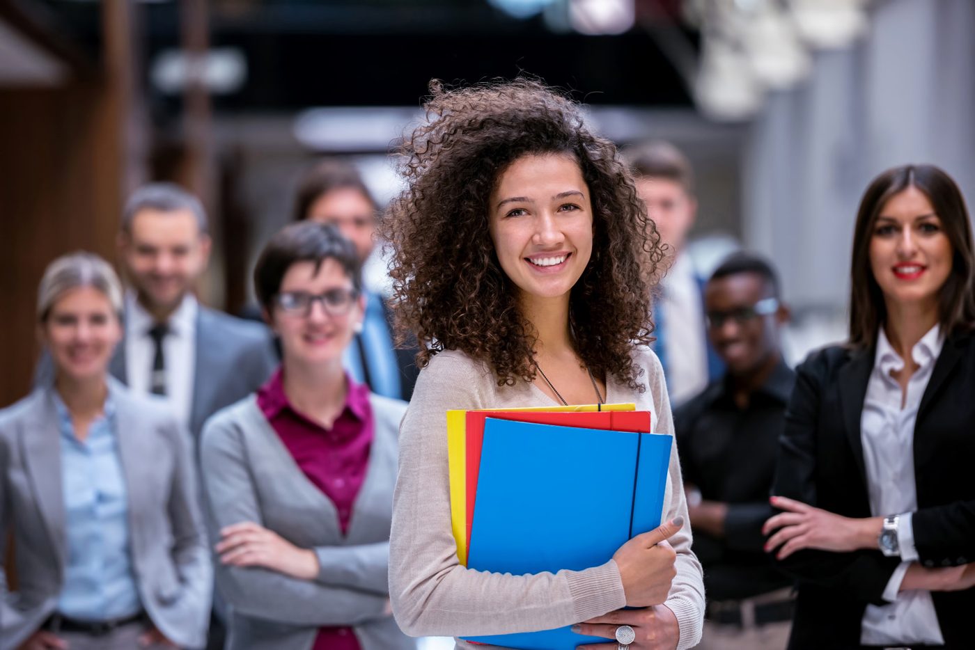 A smiling young woman holding colorful folders, with a team of professionals standing behind her